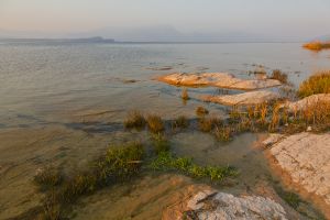 Lago di Garda. La luce del tramonto accende di toni  rossastri gli scogli in nprimo piano, che contrastano col blu del lago e dello  sfondo, reso indistinto dalla foschia.